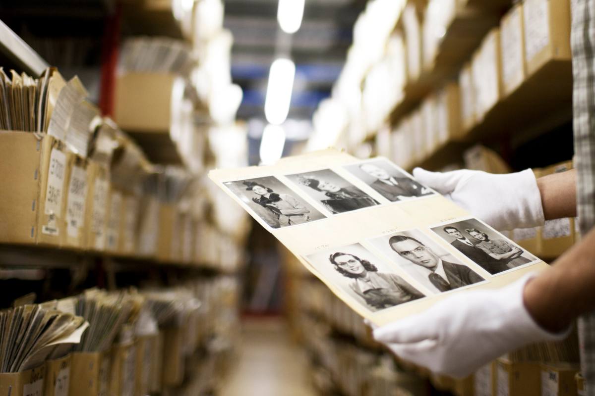 Gloved hands holding a sheet of photographs inside an archives building.